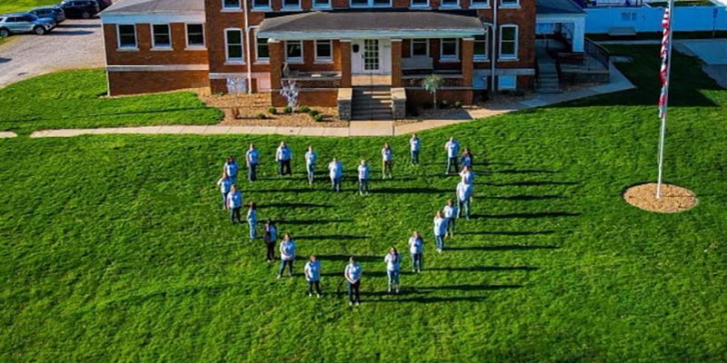 Aerial shot of a group of children standing in front of a brick building. Their bodies make the shape of a heart.